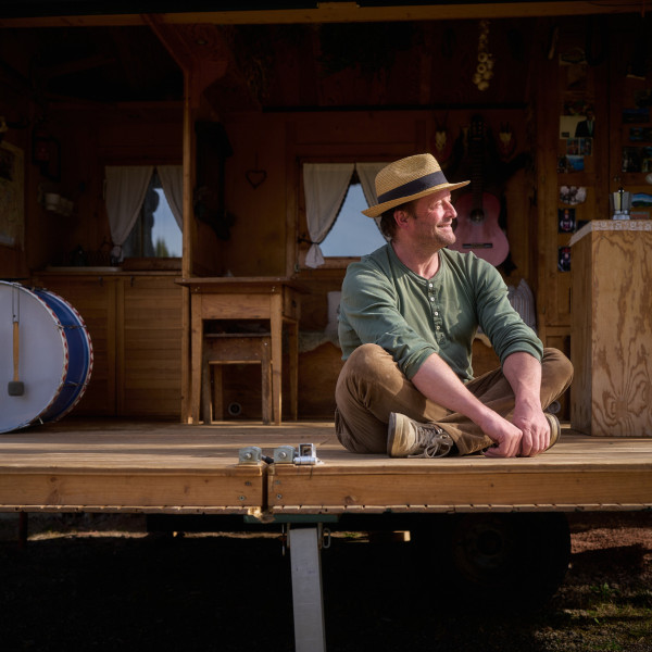 Mann mit Strohhut sitzt lächelnd auf Holzbühne eines Zirkuswagens vor rustikalem Innenraum mit Trommel und Gitarre