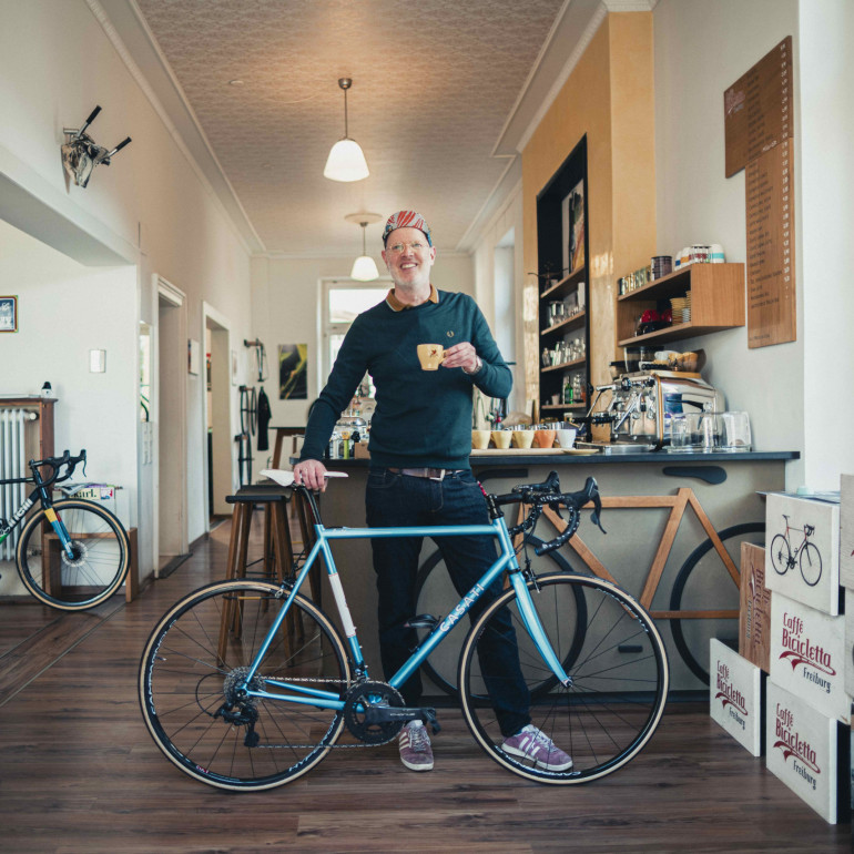 Fröhlicher Mann steht in seinem Café, vor sich ein pepsiblauen Rennrad. In der Hand hält er eine Espressotasse